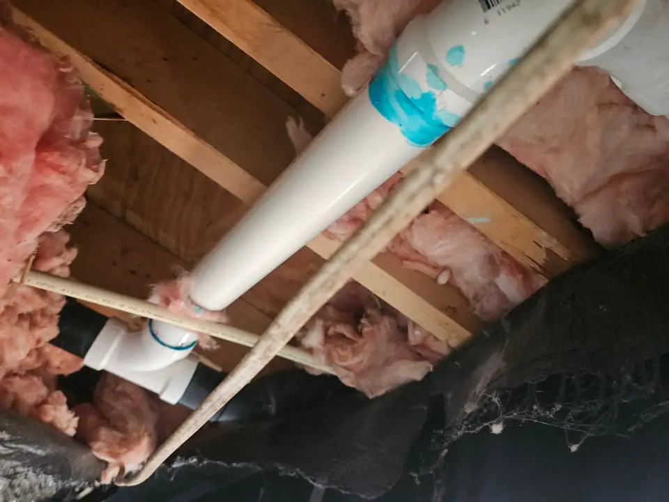 Pink insulation batts between floor joists around a PVC drain line, seen from under the home