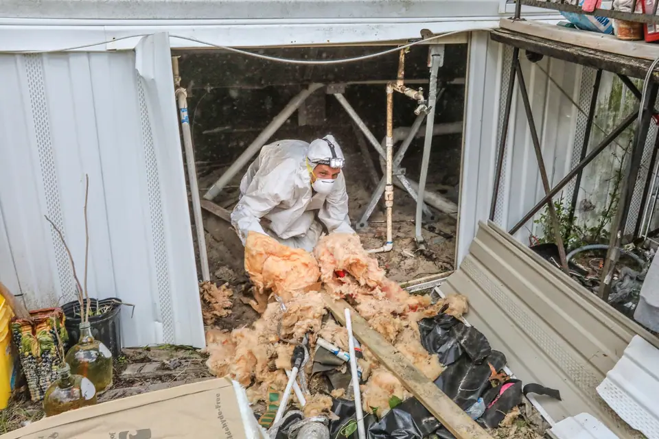 Crew member in a protective suit pulling old insulation out from under a mobile home through the skirting